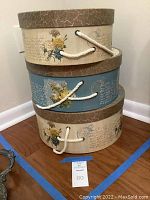 Three decorative nesting hat boxes stacked, showing beige and blue floral patterns and rope handles