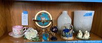 Photo of shelf with all decor items: two globes, two white vases, Lenox swans, pink floral teacup and saucer, artificial white flowers