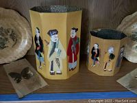 Image showing two hexagonal yellow vases hand-painted with Asian figures, with decorative woven-style plates and butterfly art nearby.