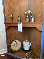 Shows wooden shelf with brass vases, silver-toned figurines, white ceramic teapot on stand, sword-shaped trivets, and Japanese framed wall art below