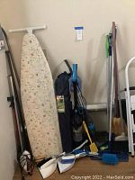 Side view of ironing board with floral cover leaning against wall with multiple cleaning tools nearby including brooms, mop, vacuum, dustpan and brush