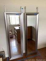 Two full-length mirrors with silver frames leaning against a wall, showing reflective surfaces and mounting hooks.