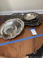 Photo showing multiple overlapping silverplate and stainless steel trays and platters placed on wooden floor near wall.