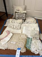 Overview photo showing folded vintage linens in whites and creams, two decorative pillows at the back, one with floral embroidery and one with humorous text embroidery.