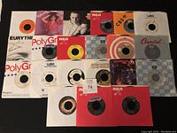 Photo showing a layout of twenty-one 45 RPM vinyl records in sleeves and loose, featuring visible labels from RCA, PolyGram, WEA, Capitol, Arista, Warner and others. Condition appears very good with some sleeves showing cover art.