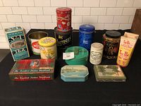Overview of 13 various vintage advertising tins and containers arranged on table against white tile background. Includes tea, yogurt, milk, honey, and biscuit tins with assorted colorful branding and artwork.