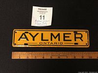 Top view of yellow metal souvenir license plate attachment with black lettering 'AYLMER ONTARIO' placed next to ruler for size reference.