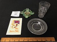 Four British collectible items displayed: commemorative glass, green glass ashtray, glass dish, and postcard. Wooden ruler used for scale.