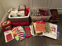 Full lot view showing stacked plastic tubs with ornaments, various Christmas gift bags laid out in front, tins, and decorative fabric rolls.