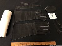 Black leather pieces cut in glove shapes spread out on black surface with ruler for scale, including fingers and palm sections along with additional smaller pattern pieces and a rolled white paper.