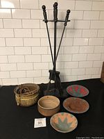 Full lot showing fireplace tools, brass pot, ceramic pot, and three handwoven baskets on black surface against white tiled wall