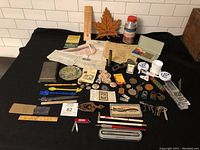 Photo of entire lot showing all items spread out on black surface against white tiled background, including skeleton keys, pens, thermometer, cups, pins, swizzle sticks, and other miscellaneous collectibles.