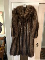 Front full length view of the brown raccoon fur coat hanging on a door, showing overall length and texture of the fur.