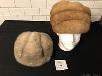 Photo showing two ladies mink fur hats side by side on a black fabric background; one beige hat lying flat and one reddish-brown hat displayed on a white mannequin head.