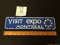 Front view of the blue metal license plate attachment with white text 'VISIT EXPO MONTRÉAL' and emblem, ruler for size reference.