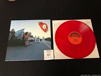 Red translucent vinyl record and cover sleeve with lyrics sheet, showing front artwork with road scene and people.