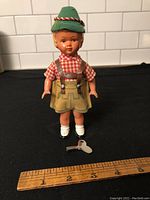 Front view of the vintage celluloid wind-up doll wearing green hat, red checkered shirt, beige skirt, white shoes, and posed standing with wind-up key in front