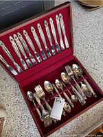 Open red velvet lined wooden case holding full set of silver-plated silverware including knives, forks and spoons arranged in ranks.