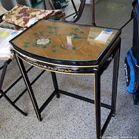 Front angled view of black lacquered side table with gold floral details and floral painting on glass top.