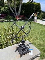 Black metal armillary sphere sculpture placed outdoors on a stone ledge, showing front and lateral views. Background includes greenery, grass, and part of a house.