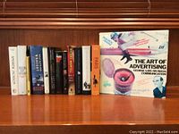 A group of twelve books arranged upright on a wooden shelf, showing a range of titles including historical and historical fiction themes along with a distinct book titled 'The Art of Advertising' on the right end.