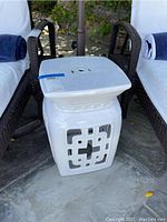 White ceramic outdoor side table/stool positioned between two lounge chairs. Shows geometric cut-out design on sides and smooth square top with ventilation holes.