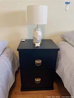 Front view of the black filing cabinet showing two drawers with metal handles and a white lamp on top between two beds.