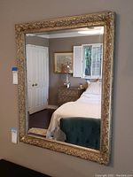Front view of rectangular gold framed mirror reflecting a bedroom area, showing ornate detailed frame and clear glass surface.