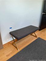 Full view of gray upholstered bench with gold-tone metal X-shaped legs on hardwood floor near a white wall and dark rug.