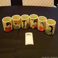 Front view of six cylindrical Japanese tea cups featuring colorful geisha designs arranged in a row on a black surface.