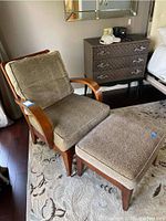 Full view of leopard print upholstered chair and ottoman set with wood frames, placed on a light-colored patterned rug, beside a dresser and bed.