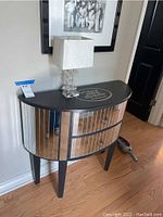 Front view of a demi-lune occasional table with mirrored drawer fronts and a decorative black top. Lamp placed on top.