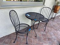 Full view of black metal outdoor bistro set with table and two chairs on tiled patio against exterior wall.