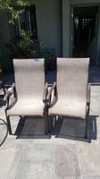 Front view of two outdoor patio chairs placed side by side on a stone patio under sunlight, showing tan mesh fabric seating and dark brown metal frames