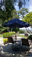 Outdoor patio umbrella fully opened with navy blue canopy and heavy metal base, situated on stone patio near cushioned chairs and small tables.