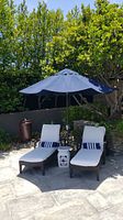 Outdoor patio umbrella with navy blue canopy, shown open above two chaise lounge chairs and a small side table on patio.