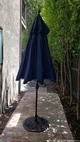 Full closed view of the outdoor patio umbrella with dark blue fabric and brown pole surrounded by trees and bricks.