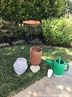 Photo showing six terracotta pots stacked, a concrete planter shaped like a face, heart-shaped stone planter, green plastic watering can, and a metal stand with terracotta bird bath bowl on top.