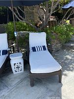 Outdoor poolside setting with one brown wicker chaise lounge chair featuring white cushions and striped pillow, small white side table visible beside chair.