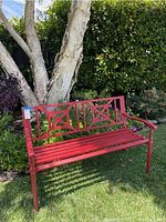 Full view of red metal outdoor bench placed on grass by a tree and garden plants, showing decorative X-pattern on backrest and slatted seat.