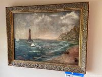 Front view of vintage framed painting depicting a seascape with lighthouse, sailboats, cliffs, beach huts.