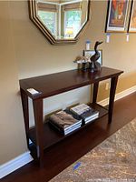 Front view of the dark wood console table with items on top and books on the lower shelf against a beige wall with a mirror above.