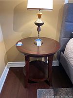 Side view of medium-sized round wooden side table with a brass and wood lamp stand on top, placed next to a grey upholstered bed and corner wall.