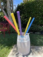 Front view of a woven cotton basket outdoors containing five foam pool noodles in red, purple, yellow, and blue colors.
