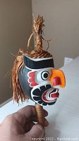 Full view of carved and painted wooden rattle with natural fiber adornment