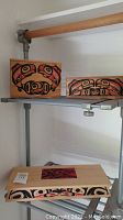 Three native art boxes on a metal shelf with wood rod above.