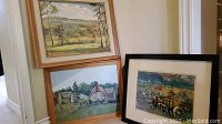 Three framed paintings leaning against a wall: two wood-framed landscapes and one in a black frame