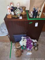 Photo showing the top of a wooden cabinet with a bronze vase, clear jar with colorful stones, pink and white faux flowers, decorative baskets, and various small decor items.