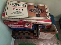Photo shows stacked boxed board games including Tripoley, Yahtzee, Password, Glass Chess, Tri-Ominos, Dream Phone, and more.