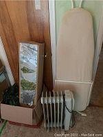 White DeLonghi oil-filled radiator heater in front of ironing board and box with Christmas decorations.